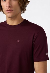 Jonge man met krullend haar die een eenvoudige bordeauxrode Champion t-shirt draagt met het logo op de borst en mouw, tegen een witte achtergrond.