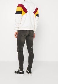 Sudadera blanca con rayas diagonales rojas, amarillas y azul marino en los hombros; vaqueros negros ajustados con salpicaduras de pintura; zapatos negros.