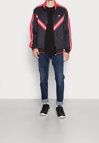 Černá sportovní bunda s červenými detaily, se V-čkovým designem, na zip a s vzorovanou texturou. Nosit s modrými džíny a černými teniskami.