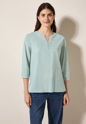 Blouse bleu clair à manches longues avec un col en V et trois boutons au niveau du col. Texture lisse, coupe légèrement ample, associée à un pantalon à rayures verticales.