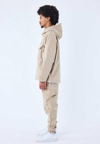 Beige lichtgewicht jas met capuchon en borstzak, gecombineerd met bijpassende beige cargobroek. De laarzen zijn wit met een textuurstof.