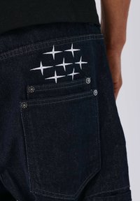 Dark denim shorts featuring a back pocket with white embroidered geometric patterns and silver-tone hardware accents.