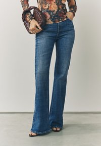 Rifľové nohavice s rozšírenými nohavicami v tmavomodrej farbe, s hladkou textúrou a klasickým päťvreckovým dizajnom, v spojení s tmavohnedou pletenou kabelkou.