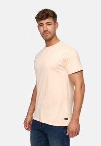 Jasnoróżowa bawełniana koszulka z krótkim rękawem, okrągłym dekoltem i małym detalem logo na klatce piersiowej. Noszona z ciemnoniebieskimi dżinsami.