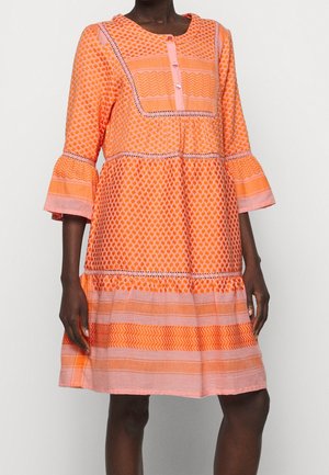 Vrouw draagt een knielange oranje en roze jurk met patroon, driekwart klokmouwen en een voorpand met knopen.