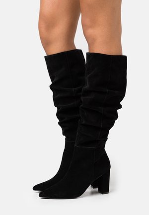 Botas de ante negras hasta la rodilla con un diseño holgado, punteras puntiagudas y tacones cuadrados. El acabado texturizado añade profundidad e interés visual.