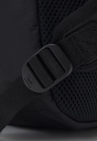 Black backpack detail featuring a mesh panel, durable fabric, an adjustable strap with a plastic buckle, and a logo embossed on the hardware.