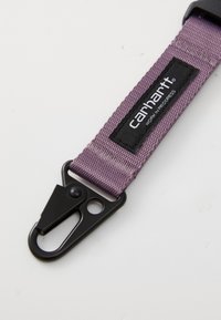 Carhartt lilla nylonrem med en sort metalclips og logo patch, med en robust, tekstureret overflade og minimalistisk design.