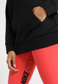 Černá mikina se zipovanou kapsou a přiléhavým střihem, kombinovaná s jasně růžovými legínami s výrazným černým logem Nike.