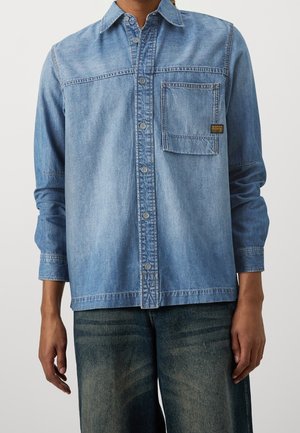 Chemise en denim bleu clair à manches longues avec fermeture à boutons et poche à rabat portée par une personne en jean foncé délavé sur fond clair.