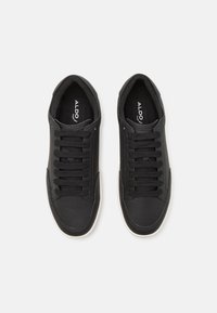Ein Paar schwarze Schnürsneaker mit durchlochteter Oberfläche und weißen Sohlen, von oben auf einem weißen Hintergrund betrachtet.