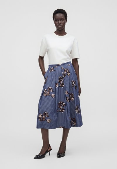 Femme portant un haut blanc à manches courtes, une jupe midi bleue à rayures verticales et imprimés floraux, et des talons slingback noirs, debout les mains dans les poches.