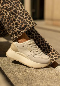 Beżowe sneakersy z teksturowanej skóry i kontrastującymi brązowymi akcentami. Gruba, biała gumowa podeszwa z rowkowanym wzorem. Noszone z spodniami w leopardzi wzór.