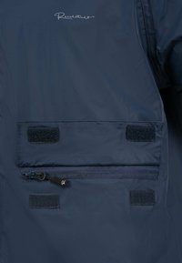 Marineblauwe jas met een gladde textuur, voorzien van een ritszak en klittenbandsluitingen. Logo weergegeven in de bovenhoek.