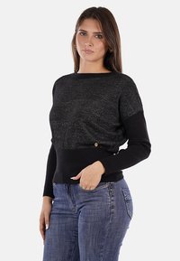 Maglione nero con sezione superiore testurizzata e sezione inferiore liscia a costine, dotato di un delicato bottone di accento. Indossato con jeans blu.