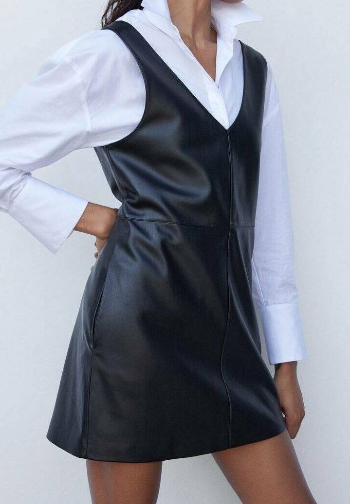 Femme portant une robe en cuir noire sans manches par-dessus une chemise blanche à manches longues avec un large col, posant avec une main sur la hanche.