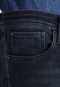 Jeans de mezclilla oscura con un bolsillo frontal visible, con remaches de metal y costuras en contraste a lo largo de los bordes. Tejido denso y textura suave.