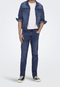 Donkerblauwe spijkerjas met vervaagde accenten, witte T-shirt eronder, gecombineerd met bijpassende blauwe spijkerbroek en zwarte sneakers.