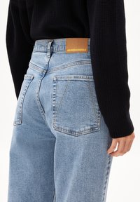 Jean en denim bleu clair avec taille haute, cinq poches, couture visible et une étiquette en cuir marron portant l'inscription "ARMEDANGELS" au dos.