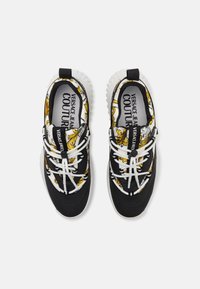 Baskets à motifs noir et jaune avec une semelle blanche, des lacets élastiques et un logo sur la languette, dotées d'un dessus en tissu lisse.