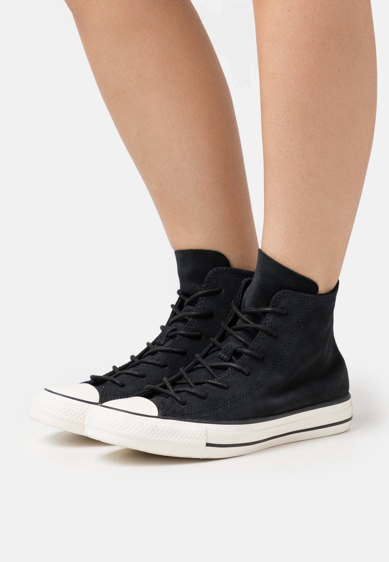 Jambes portant des baskets montantes noires avec semelles et bouts en caoutchouc blanc, présentées sur un fond clair uni.
