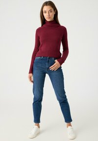 Suéter de cuello alto burdeos, textura acanalada, diseño ajustado, combinado con jeans azul oscuro y zapatillas blancas. Conjunto sencillo y casual.