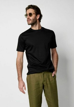 Man met ronde zonnebril, zwart T-shirt met korte mouwen en olijfgroene broek, staand met één hand in zijn zak, kijkt naar links.