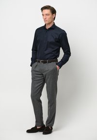 Marineblauw overhemd met knopen, grijze textuurbroek, bruine riem en donkerleren moccasinschoenen. Model staat met de handen in de zakken.