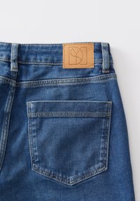 Blauwe spijkerbroek van denim met een leren label waarop een logo staat. De spijkerbroek heeft een klassiek vijf-pocket design en zichtbare stiksels.
