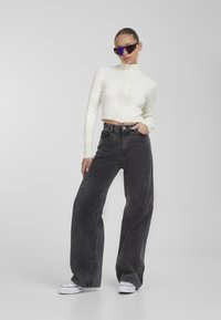 Cropped white ribbed turtleneck sweater paired with high-waisted, wide-leg black jeans and white platform sneakers, accessorized with blue sunglasses.