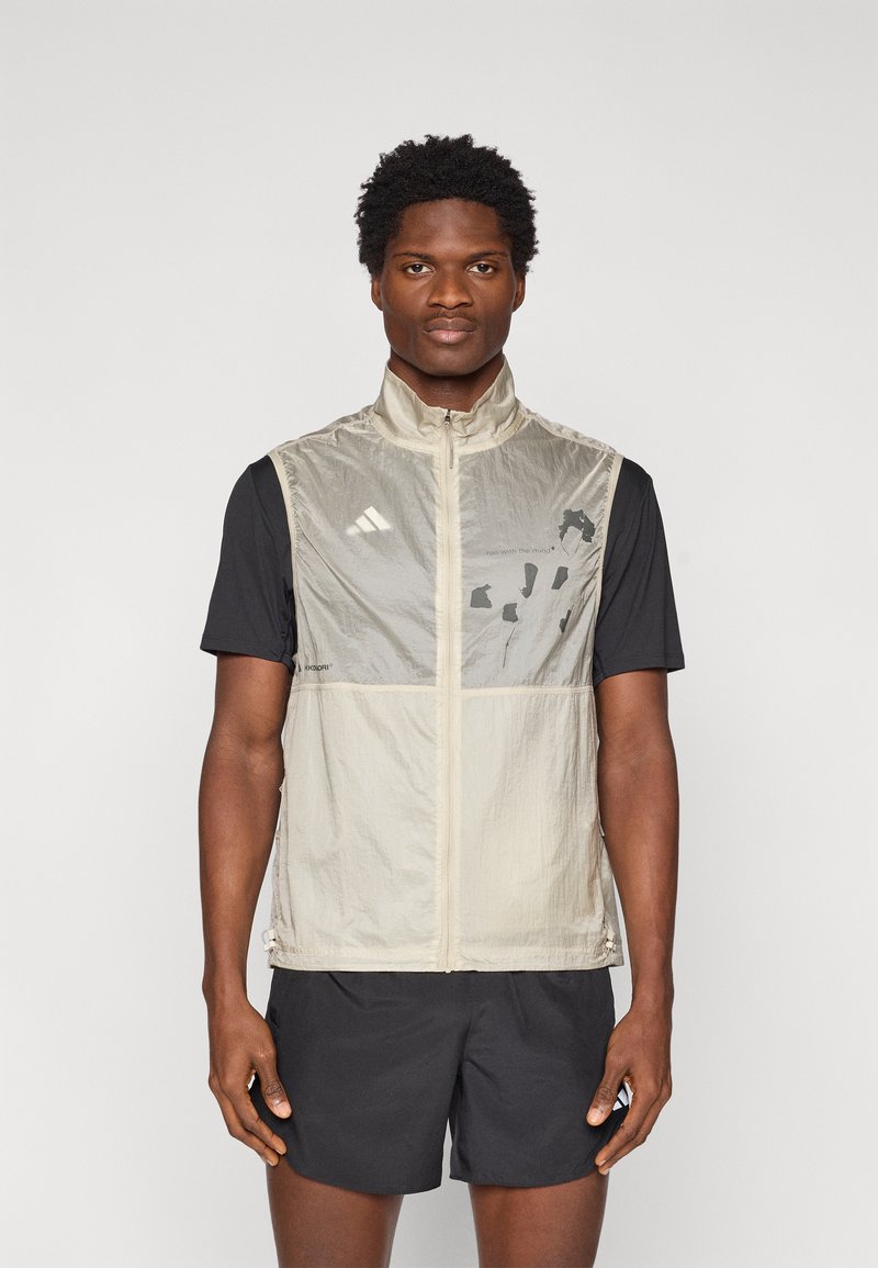 Ελαφρύ μπεζ γιλέκο χωρίς μανίκια με ψηλό γιακά, κλείσιμο με φερμουάρ και ανακλαστικές λεπτομέρειες. Μπροστινό μέρος με σχέδιο και μαύρο κοντομάνικο πουκάμισο από κάτω.