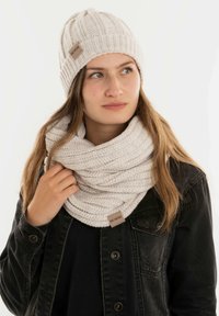 Gehaakte crèmekleurige beanie met een geribd ontwerp, gecombineerd met een bijpassende chunky infinity sjaal, beide met merklabels.