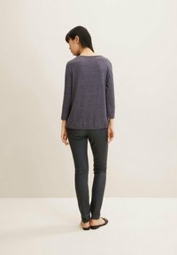 Pulover texturat violet cu mâneci până la trei sferturi, asortat cu pantaloni negri, ajustați. Modelul este întors cu spatele, evidențiind detaliile și tăietura din spate.