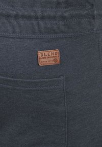 Tissu bleu foncé avec une taille côtelée et une poche. Présente une étiquette en cuir tan avec "BLEND" et "GLOBAL JEANMAKER" en relief.