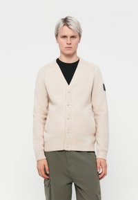Beige cardigan with a V-neck, seven buttons, ribbed cuffs and hem. Textured knit pattern with a logo patch on the left sleeve.