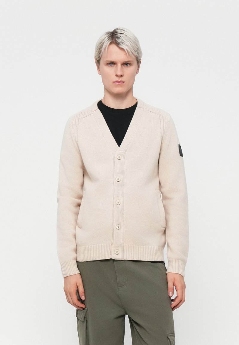 Beige Cardigan mit V-Ausschnitt, sieben Knöpfen, gerippten Bündchen und Saum. Strukturierter Strickmuster mit einem Logo-Patch am linken Ärmel.