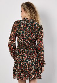 Blumenkleid mit dunkelgrünem Hintergrund und einem Muster aus roten und weißen Blumen, gesmokter Taille und geschichteten Rüschen am Saum. Lange Ärmel.