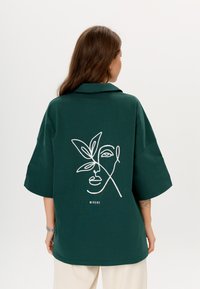 Donkergroen oversized shirt met een kraag. De achterkant heeft witte lijntekening van een gezicht met een blad, en daaronder de tekst "MIRONS". Gladde textuur.