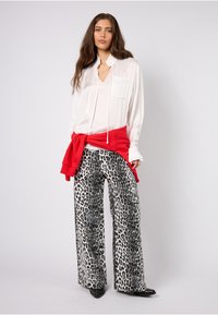 Hvit bluse med lange ermer, krage og lomme, kombinert med svart-hvite leopardmønstrede vide bukser, fremhevet med en rød genser knyttet rundt livet.