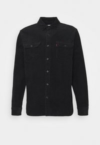 Camisa de pana negra con collar tipo botón, mangas largas, dos bolsillos en el pecho y una textura sutil; cuenta con botones negros y una pequeña etiqueta de logo.
