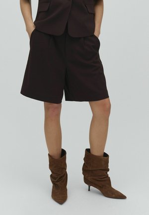 Osoba oblečená v čiernych na mieru šitých šortkách, zodpovedajúcom saku a hnedých mäkkých semišových členkových čižmách na podpätku, stojaca s rukami vreckách.