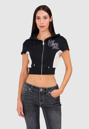 Jonge vrouw met lang donker haar die een zwart-witte cropped hoodie met rits en donkergewassen jeans draagt, staand met één hand in de zak.