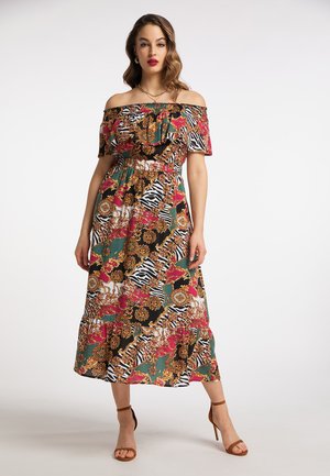 Vrouw draagt een kleurrijke off-shoulder midi-jurk met een gemengd bloem- en dierenprint, gecombineerd met bruine sandalen met hoge hakken, staand tegen een witte achtergrond.