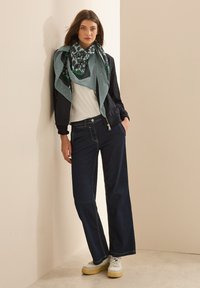 Jean large en denim bleu, t-shirt blanc, bomber bleu marine, écharpe à motifs verts et noirs, et baskets crème avec des semelles jaunes.
