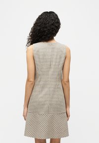 Γυναίκα με μακριά σγουρά μαλλιά φοράει αμάν ικο, καρό φόρεμα σε μπεζ και μαύρο χρώμα, στέκεται γυρισμένη προς τα πίσω μπροστά σε ένα απλό λευκό φόντο.