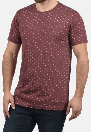 Mann trägt ein kastanienbraunes T-Shirt mit kurzen Ärmeln und kleinen weißen Tropfenmustern, kombiniert mit dunkelblauen Jeans, steht mit entspannten Händen.
