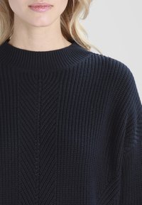 Navy knitted sweater with ribbed texture and high neckline. Features vertical chevron pattern along the front. Soft fabric.
