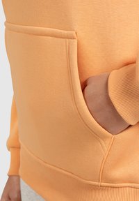 Sweat-shirt orange avec une poche kangourou, des poignets et un ourlet côtelés. Le tissu semble doux et texturé, avec des détails de couture visibles.