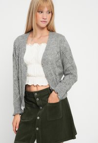 Cardigã cinza tricotado com padrão de diamante, punhos canelados e botões, combinado com um top branco cropped e uma saia de veludo verde.