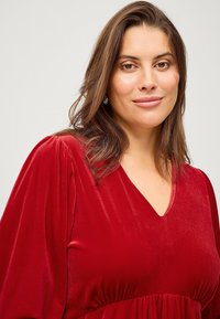 Rode fluwelen jurk met een V-hals en een gathered taille. Heeft lange mouwen en een zachte, gladde textuur.